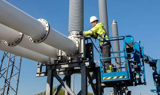 Man in yellow florescent jacket and hard helmet on a crane next to a huge tower of metal silver pipes